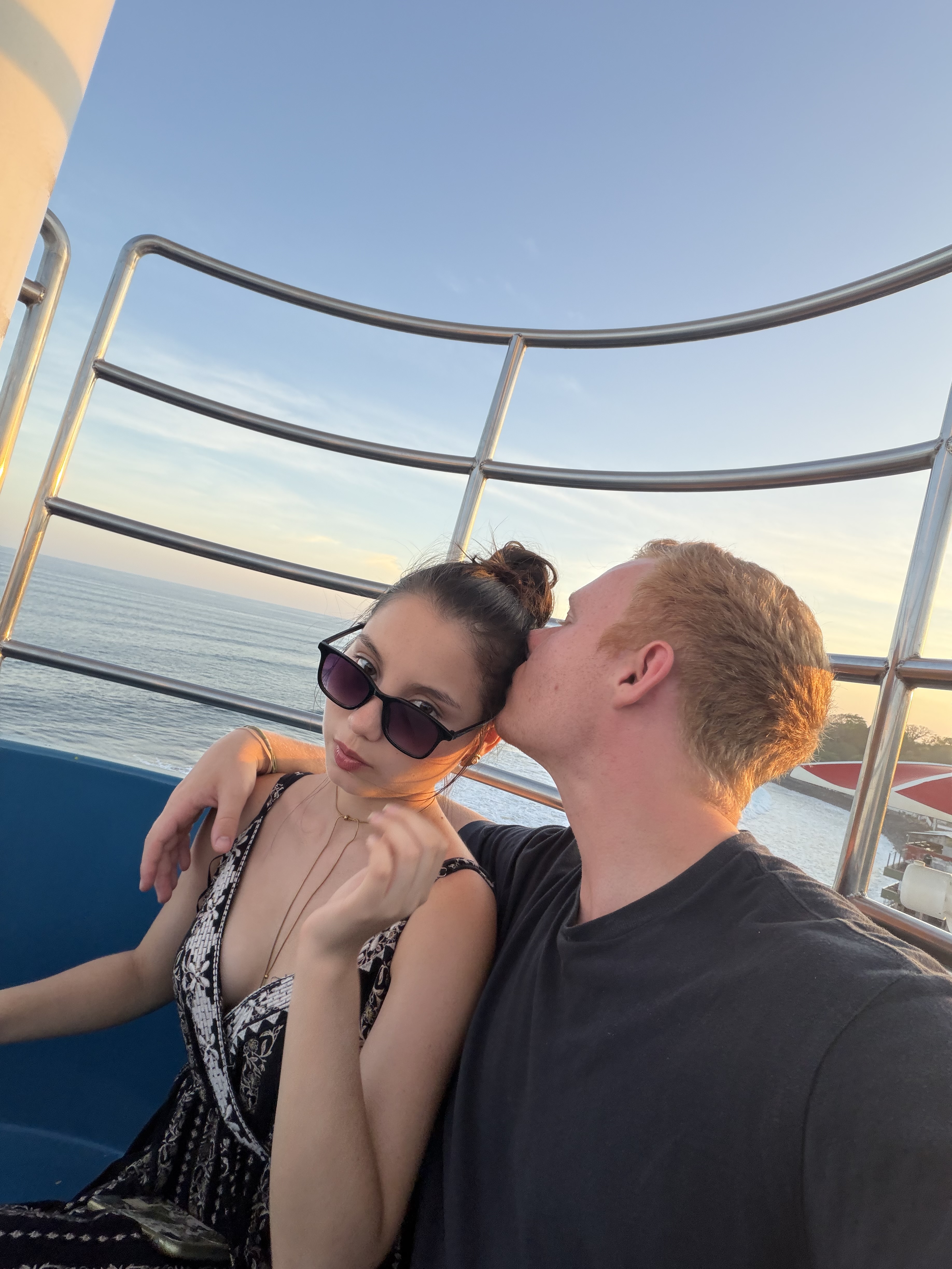 A couple on a cruise deck at sunset, one kissing the other on the head